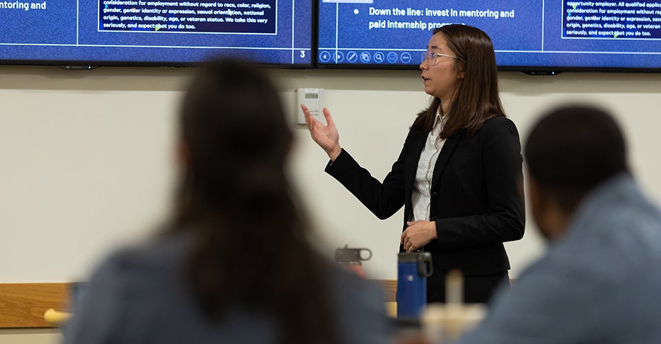 A young woman in professional dress references a slide as a panel of judges is visible in the foreground.