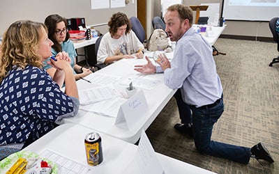 Erick Mueller on his knees in a classroom as he explains a concept to entrepreneurs seated at desks.