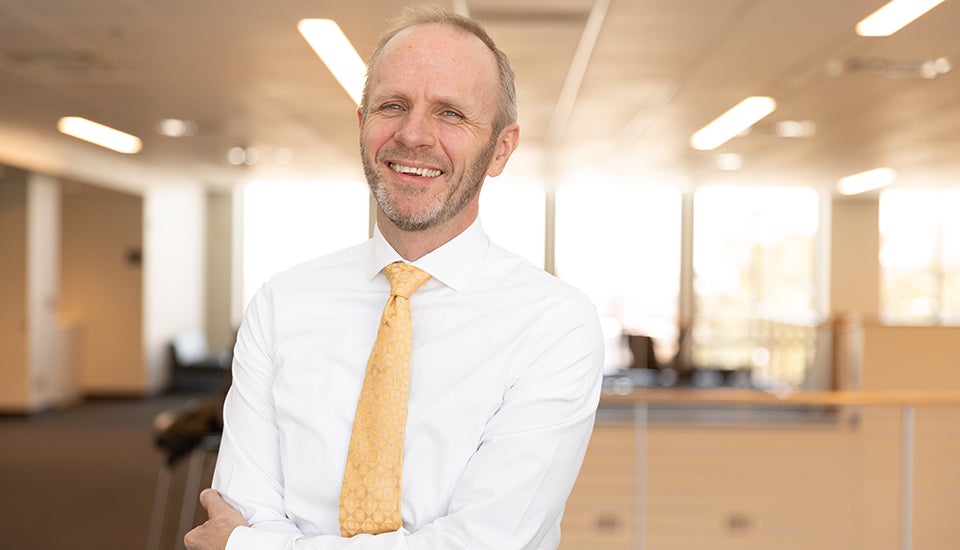 Kai Larsen in professional dress smiling with his arms crossed in an academic building. 