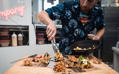 Close up of waffles on kitchen counter. James Fogerty is visible in the background as he adds toppings. 