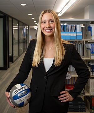 Taylor Simpson poses in the business library in professional attire while holding a volleyball.