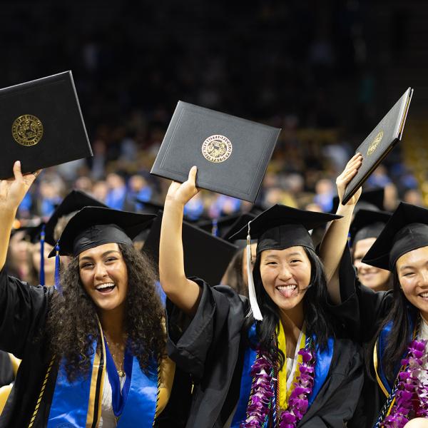 A group of graduates smiles while holding their diplomas in the air.