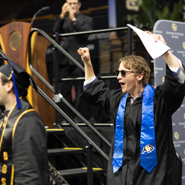 A graduate wearing sunglasses raises their arms in the air.