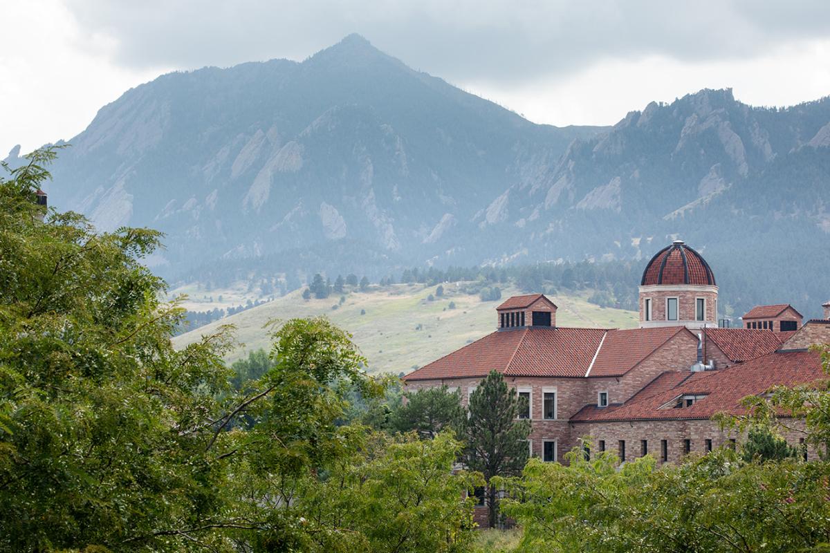 Photo of the Koelbel Building from a distance with the Flatirons in the background and foliage in the foreground.