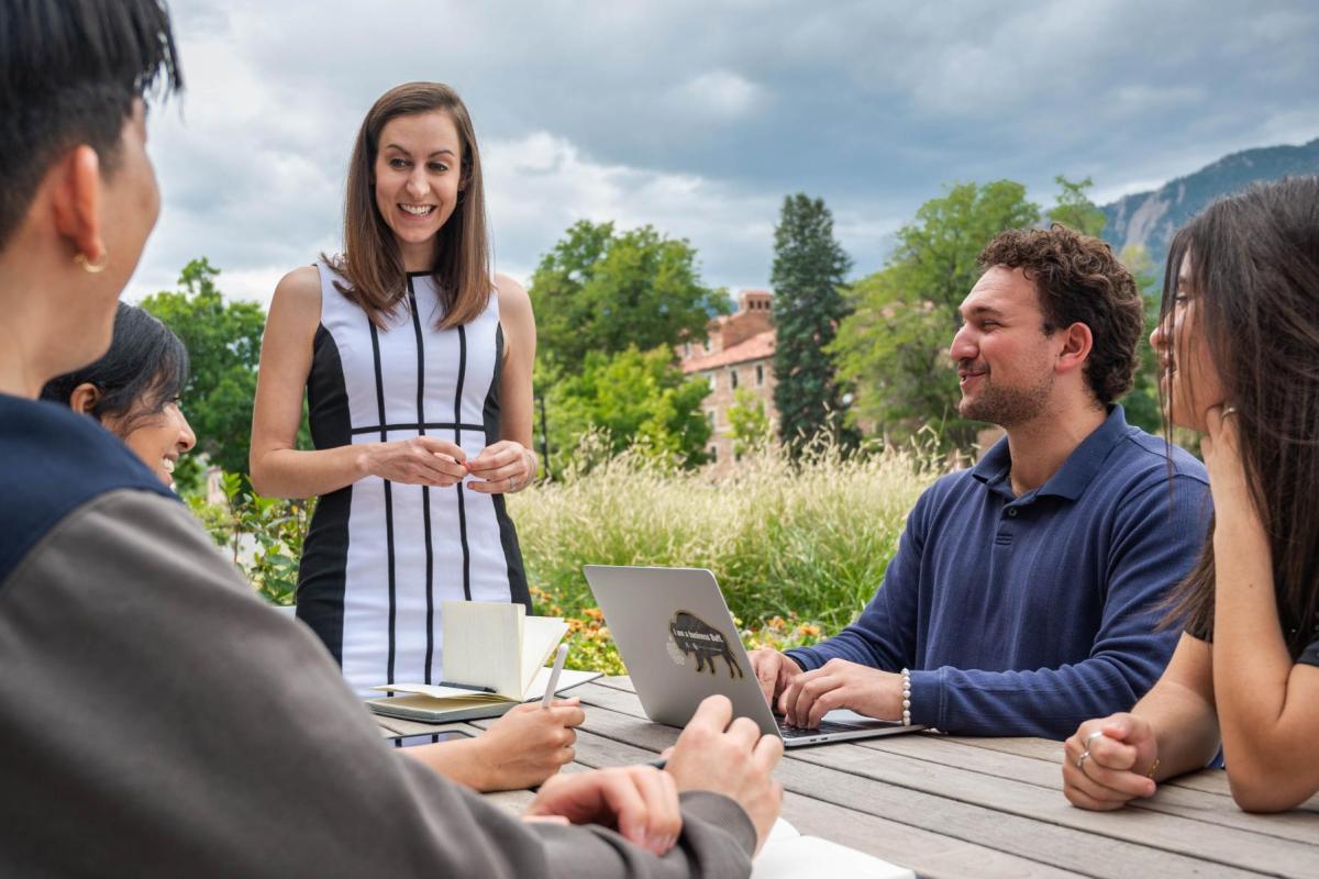 A professor speaks with students at an outdoor table.