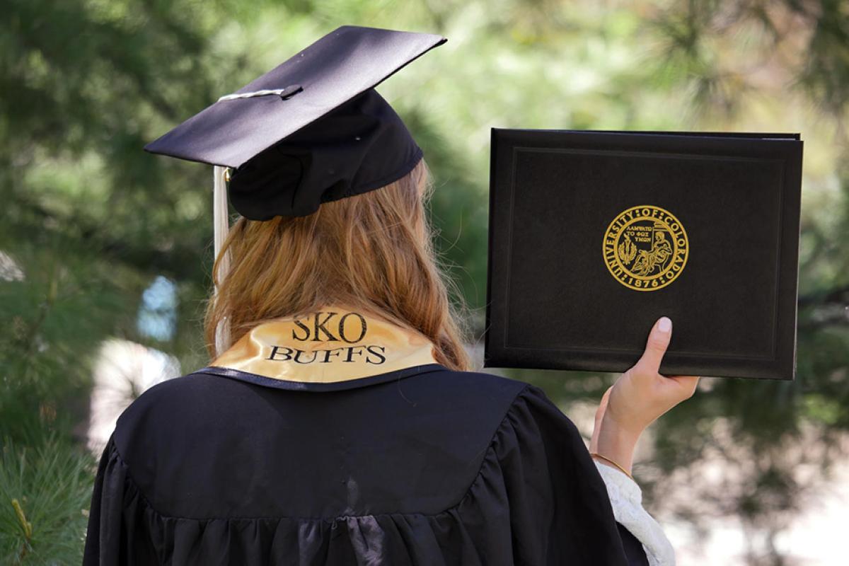 A student in her cap and gown, holding her diploma, seen from behind.
