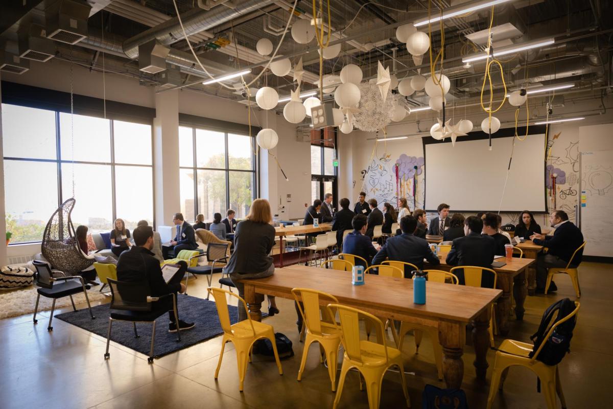 Students in professional attire gather in a modern space to listen to a guest speaker.