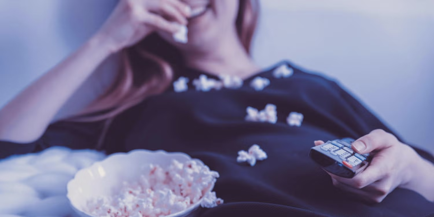 A woman eats popcorn while sitting on the couch holding a remote