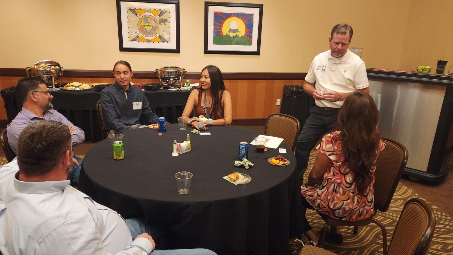 Five people sitting around a round table with a sixth person standing talking to someone seat at the table