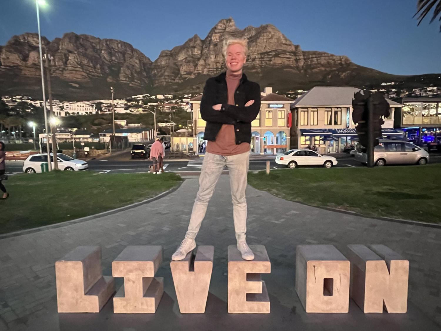 Alex Ayala standing in from of the mountains in Camps Bay, South Africa, on top of a "Live on" sign