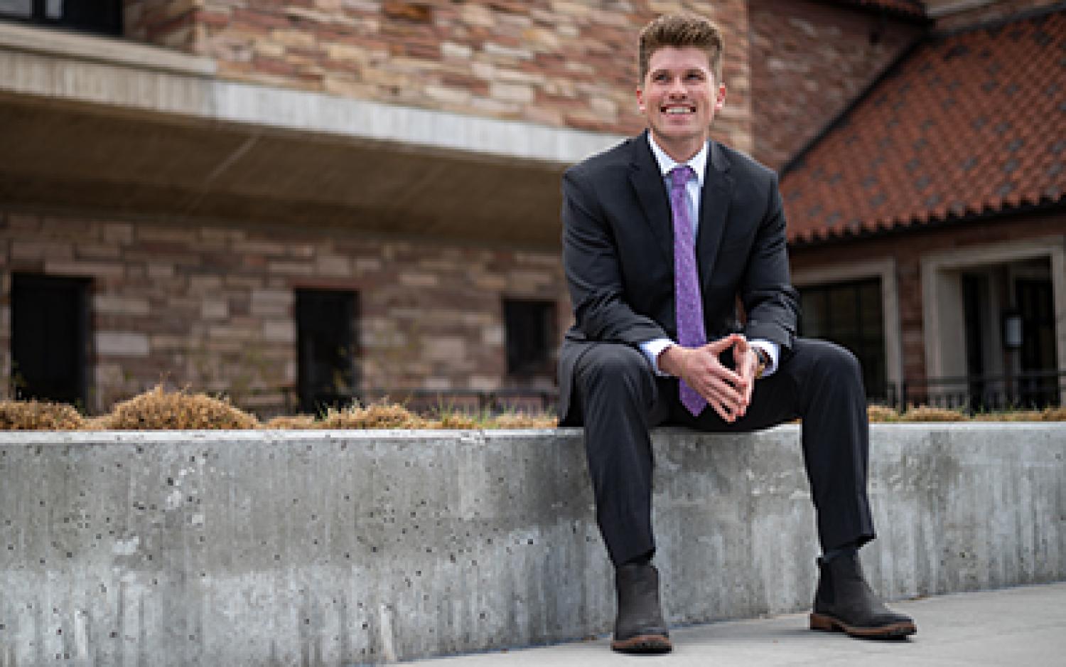 Andy Stasch smiles while wearing a suit and sitting outside of the Rustandy Building.