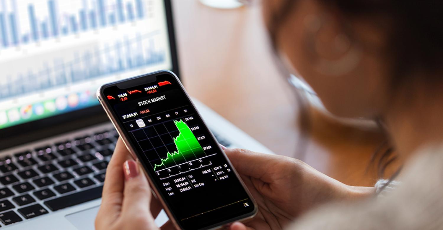 A woman looks at stock market data on a smartphone. In the background is a computer terminal showing financial data.