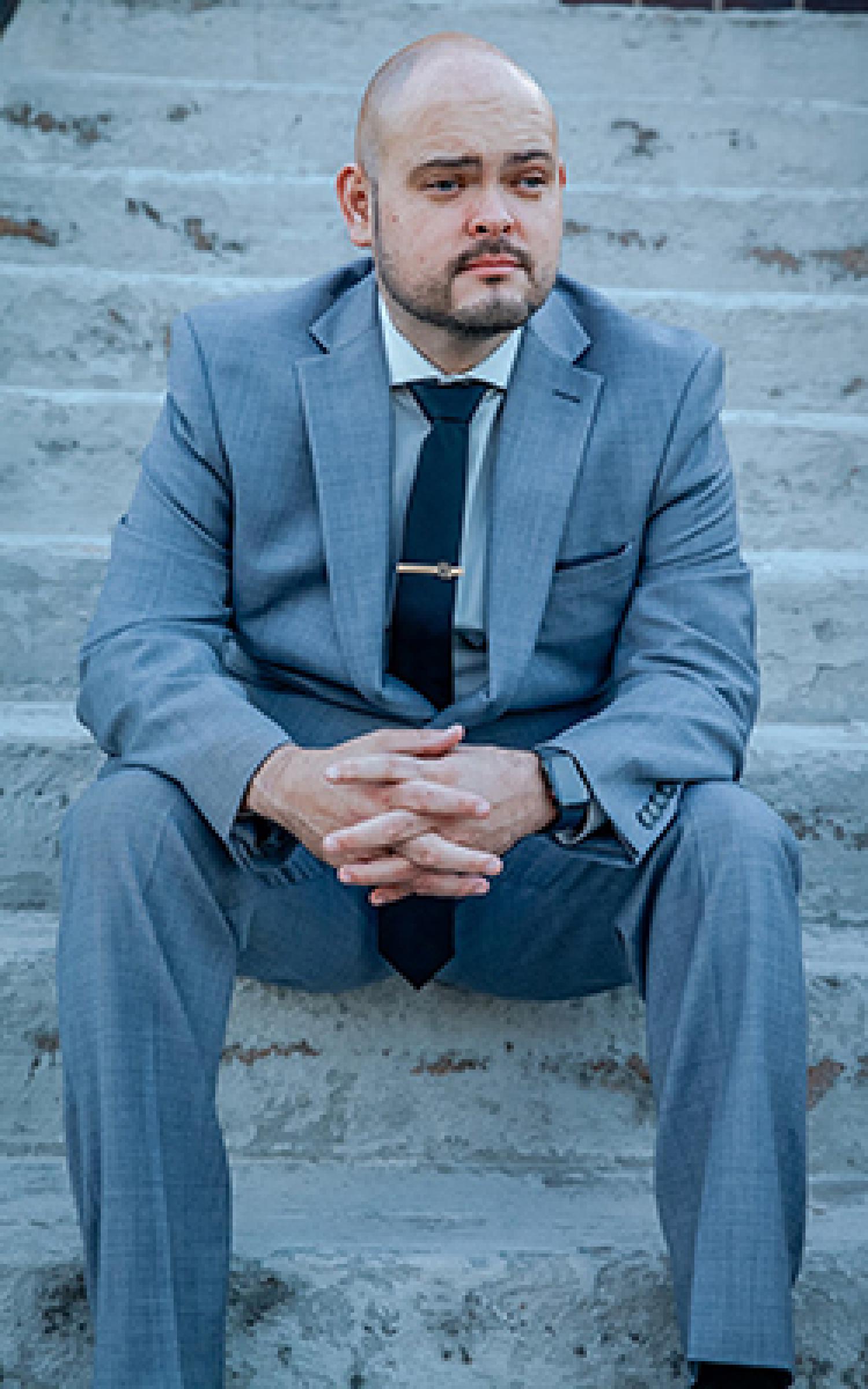Josh Ortega in a gray suit, seated on a staircase.