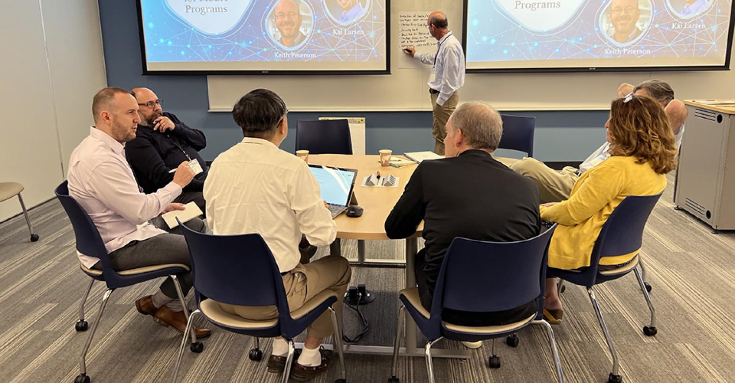 A group of professors discussing their work around a table while a colleague writes notes at a whiteboard in the background.