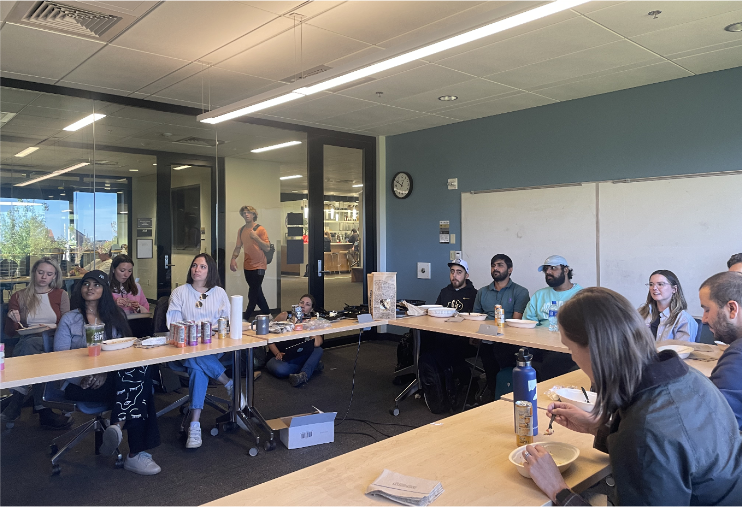 Group of students sitting at desks while eating lunch and listening to presentation