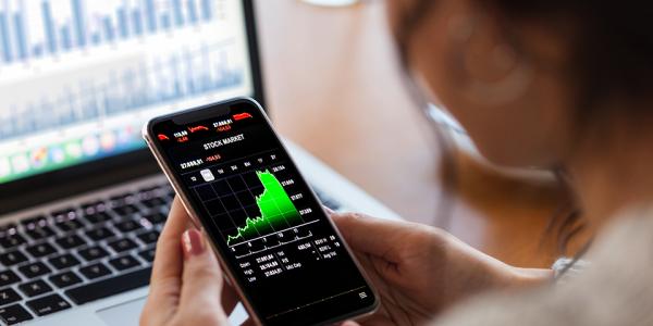 A woman looks at stock market data on a smartphone. In the background is a computer terminal showing financial data.