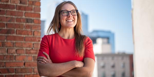 Colette, in a red dress, looks out over the Denver skyline from the roof of her building.