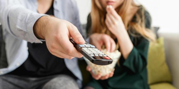 Man points a remote while sitting on a couch with a woman eating popcorn