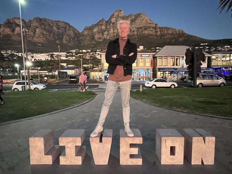 Elex Ayala standing in from of the mountains in Cape Town, South Africa, on top of a "Live on" sign