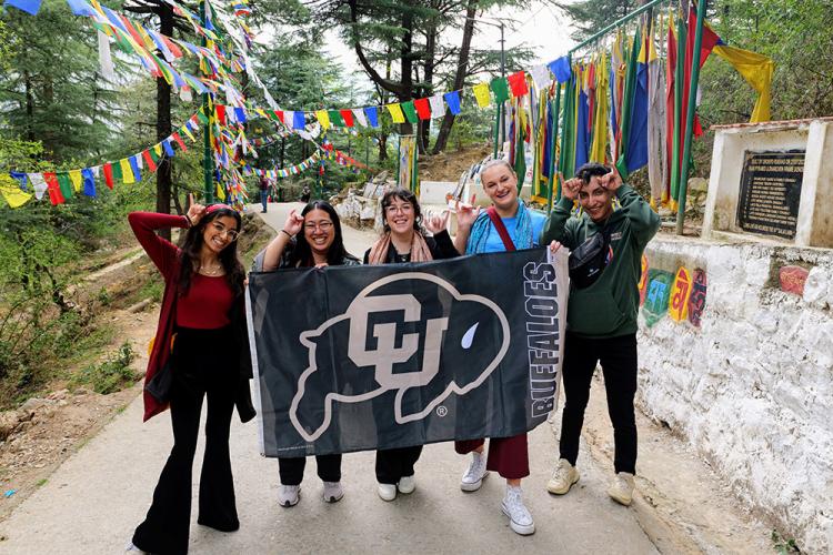 Photo of students holding a CU Boulder flag and posing with the CU Boulder hand sign