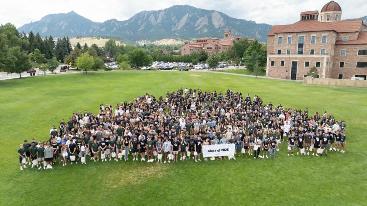 The Class of 2028 poses for a group photo on the quad with the Koelbel Building and Flatirons in the background.