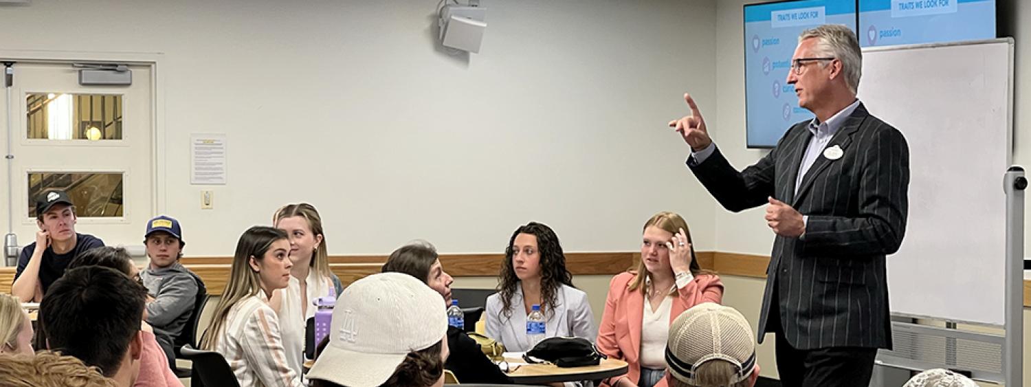 Clark Jones, in a suit, makes a point in the front of a crowded classroom. Other students look back to see the student Clark is talking to. 