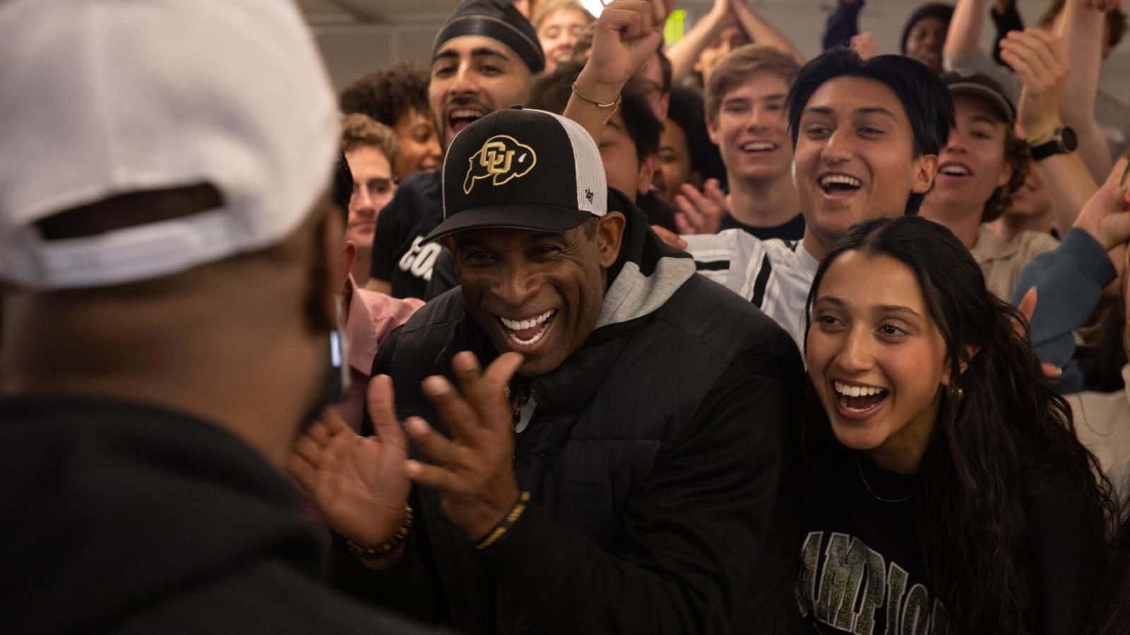 Deion Sanders laughs and claps surrounded by students during an event at Leeds