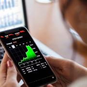 A woman looks at stock market data on a smartphone. In the background is a computer terminal showing financial data.