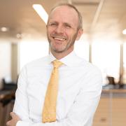 Kai Larsen in professional dress smiling with his arms crossed in an academic building. 