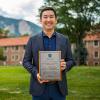 Tony Kong poses on the quad with his award. The mountains are visible in the background.