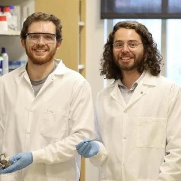 Kian Lopez (L) and Anthony Straub (R) dressed In lab coats and gloves in the lab.