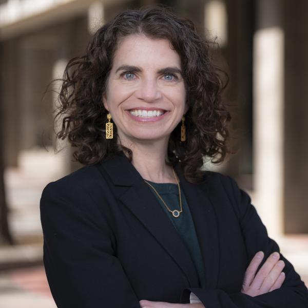 Amy Javernick-Will wearing a black blazer, earrings and a necklace in front of a blurred building