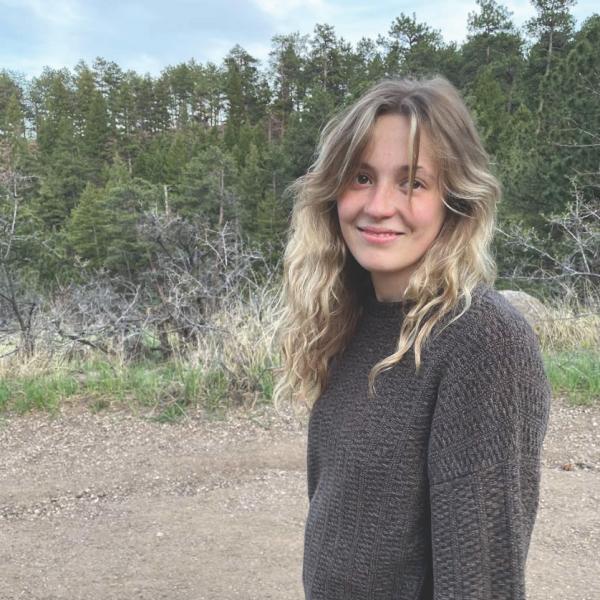 Macy Will standing in a sweater with some pine trees in the background.