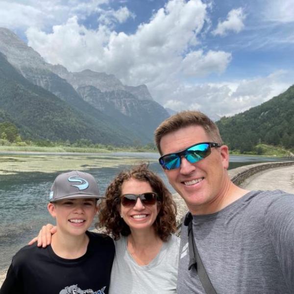 Amy Javernick-Will and her teenage child and husband casually dressed with a scenic mountain landscape and a river in the background. The father and son are wearing sunglasses and Amy has curly hair.