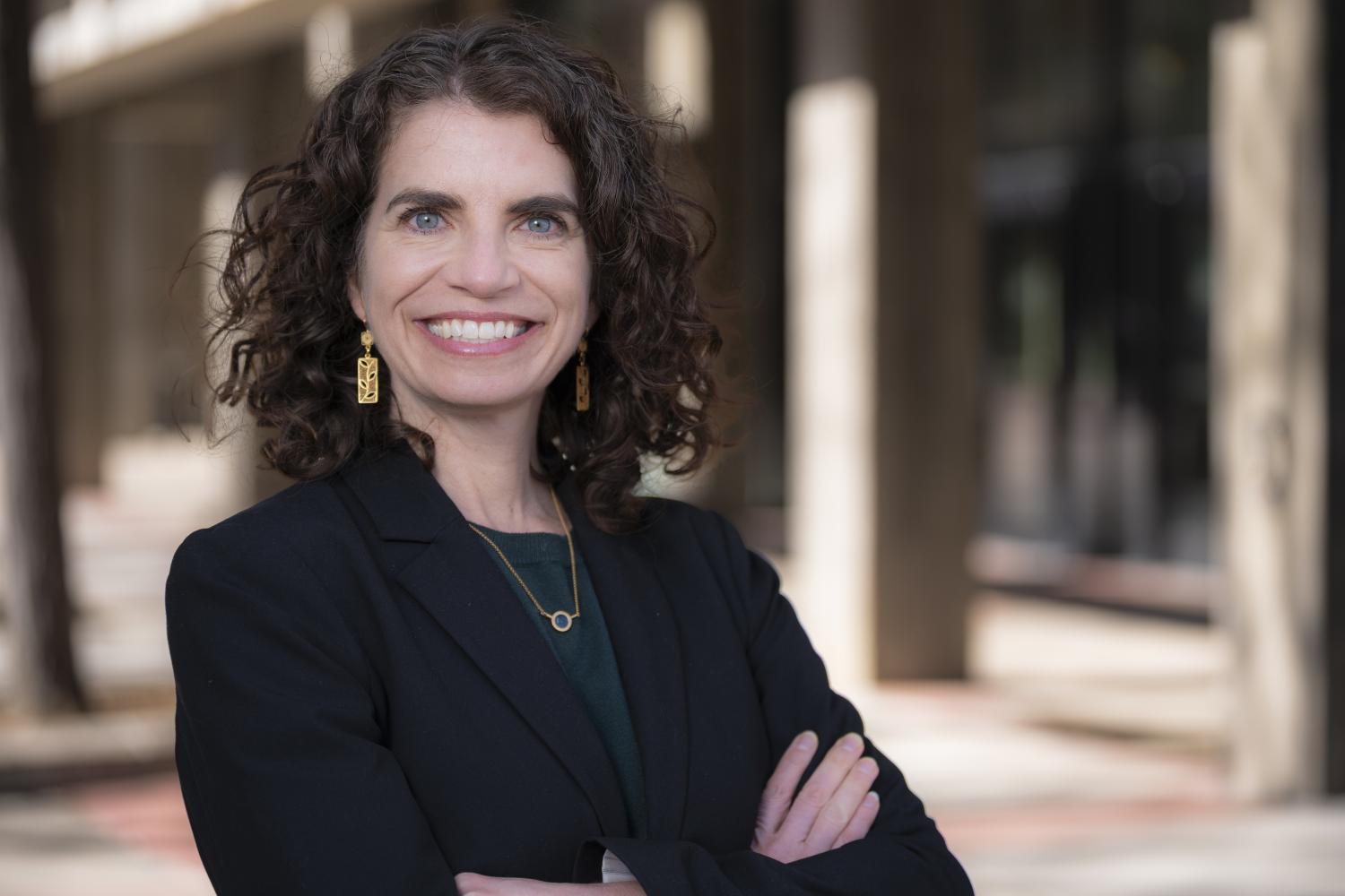 Amy Javernick-Will wearing a black blazer, earrings and a necklace in front of a blurred building