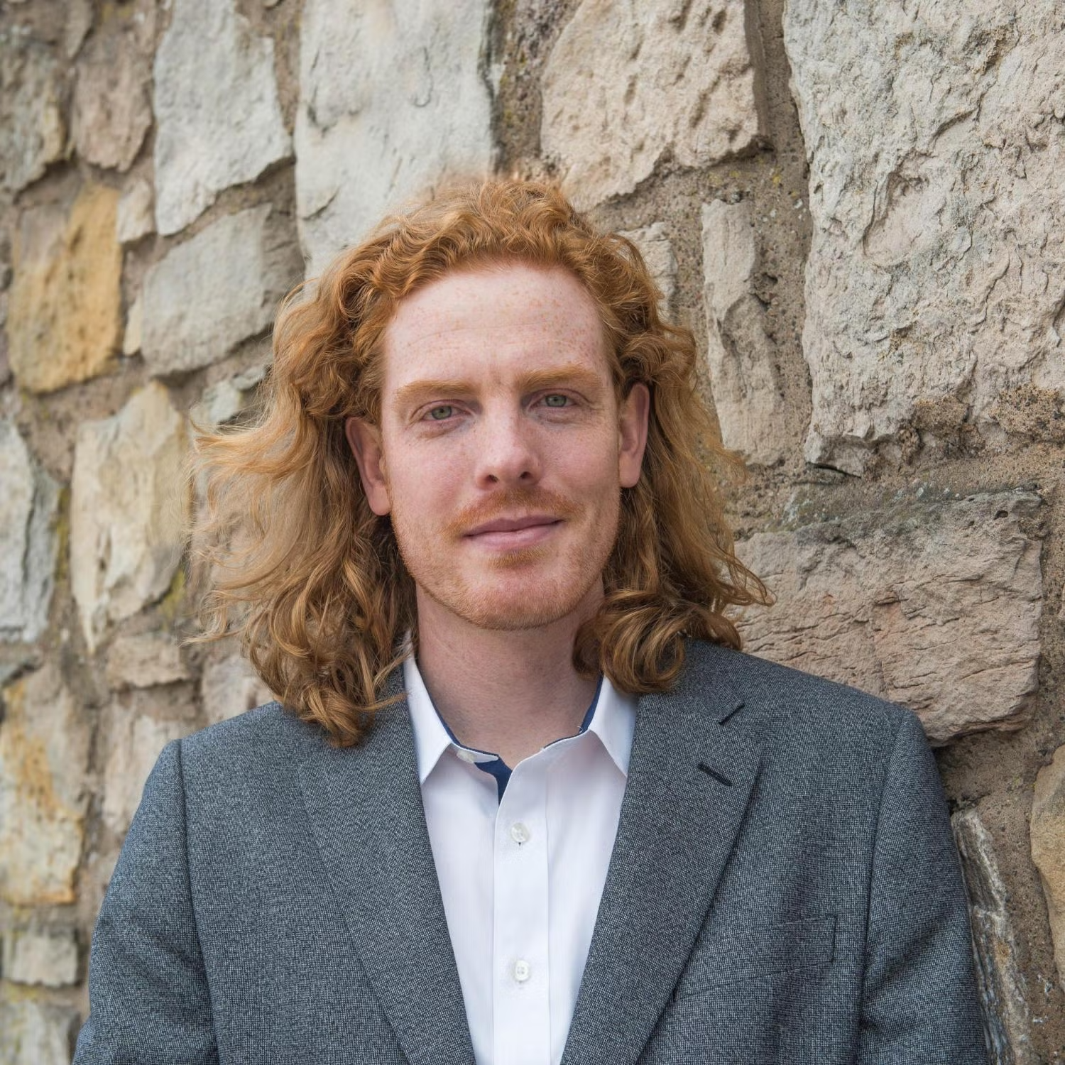 Jay Arehart in a white shirt and gray blazer in front of a stone wall.