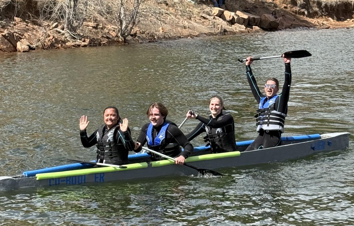 Four members of the Concrete Canoe team in wet suits sit in the concrete canoe on the water with the person in the back raising their paddle high.