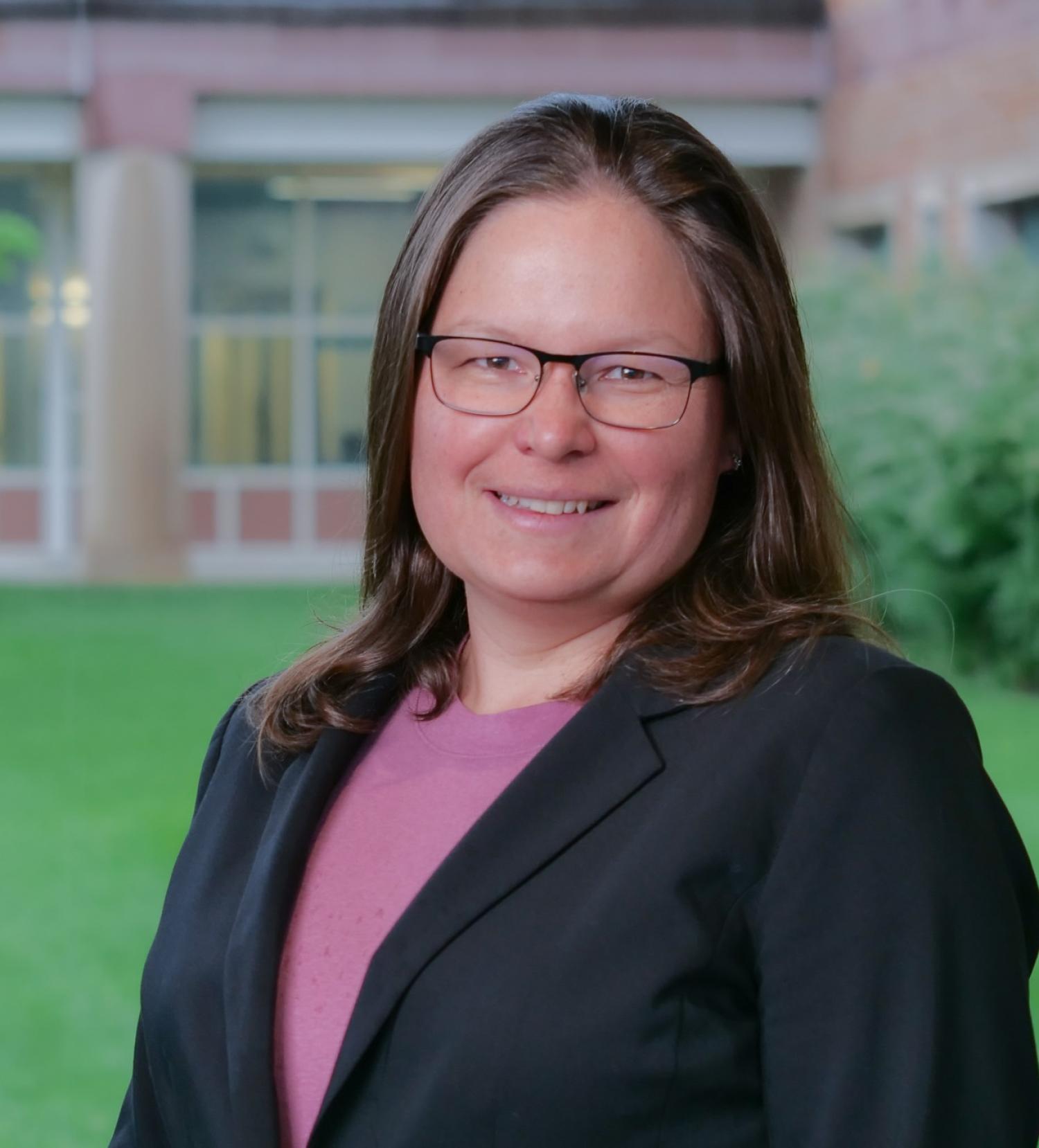 Mija Hubler with glasses on in a suit jacket and pink top in front of grass and behind that a blurred part of the SeeC building