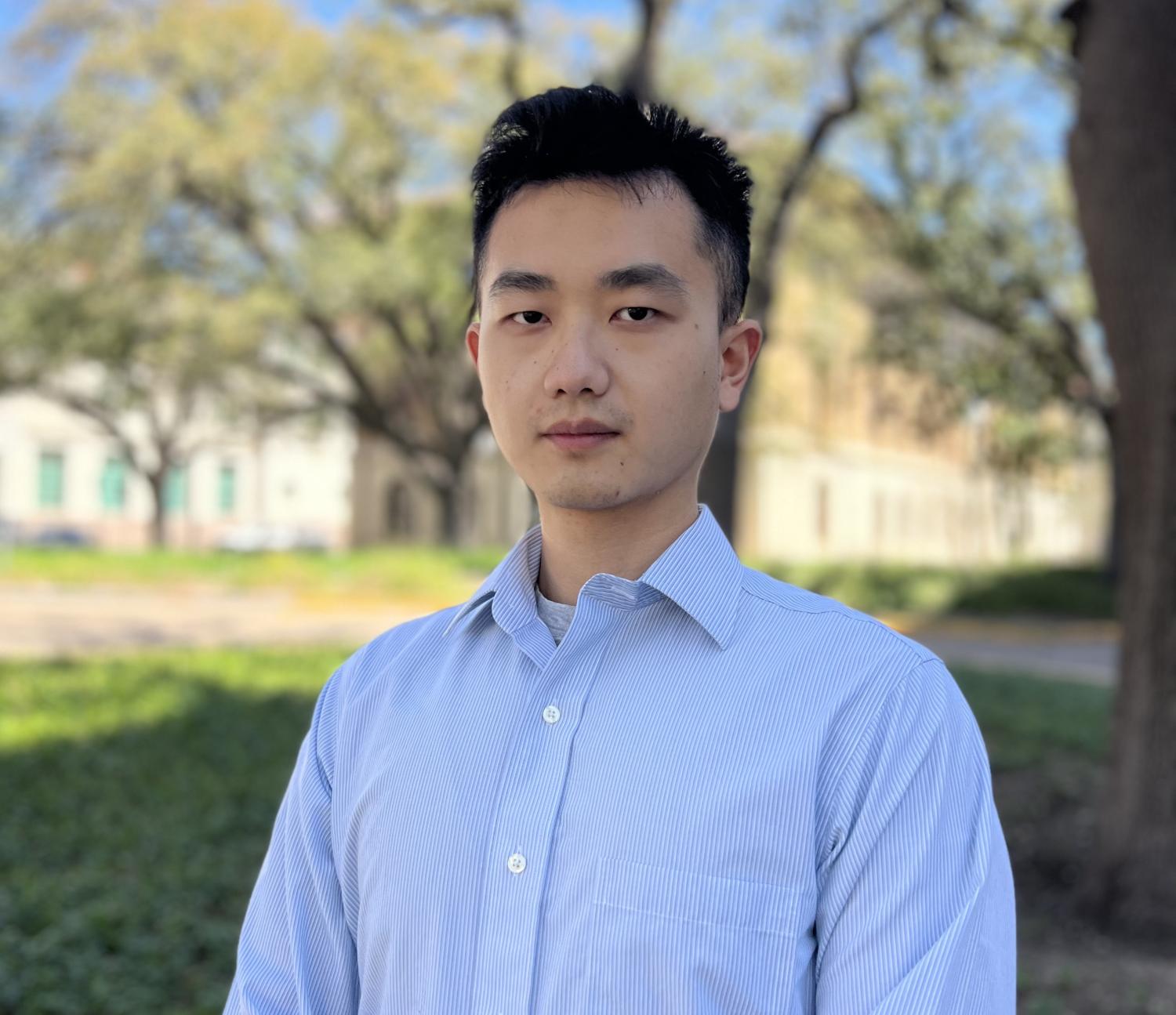 Zhi Li in a button-down shirt with some trees in the background and a building behind the trees.