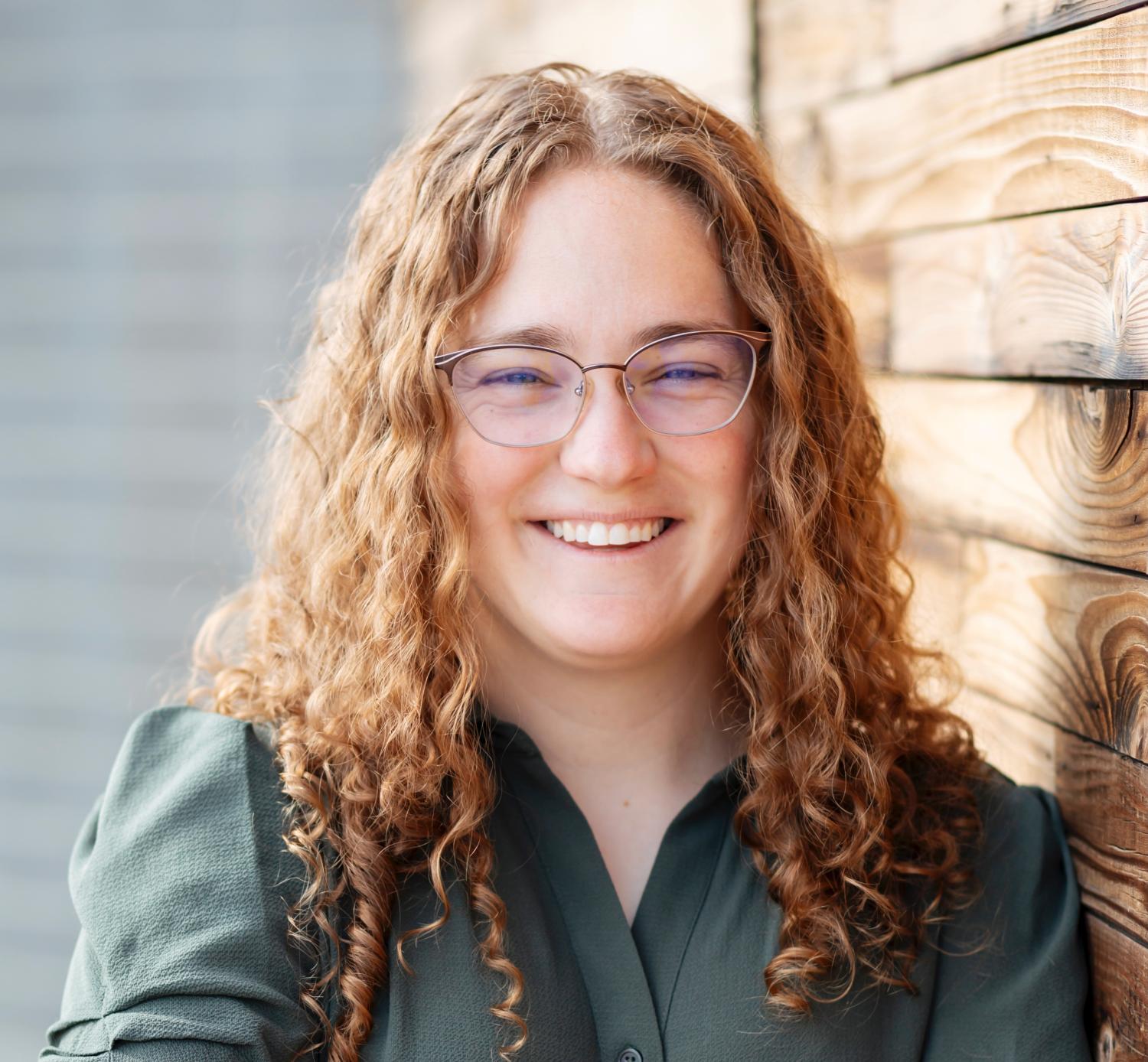 Laura Sunberg wearing glasses and red curly hair beneath her shoulders in a casual green shirt.