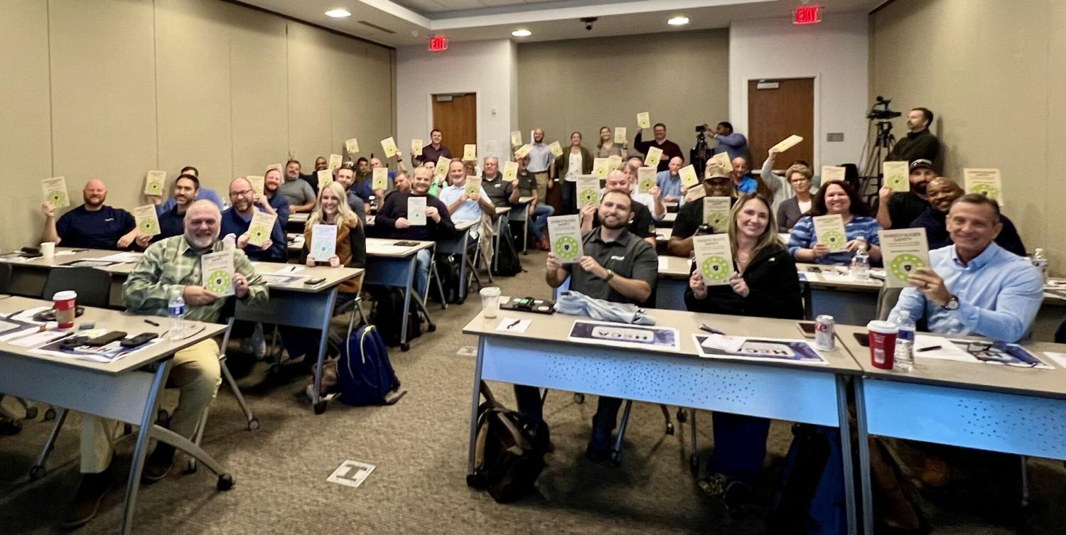 About 30 industry members of the Construction Safety Research Alliance sit at desks while proudly holding up Matt Hallowell&rsquo;s new book.