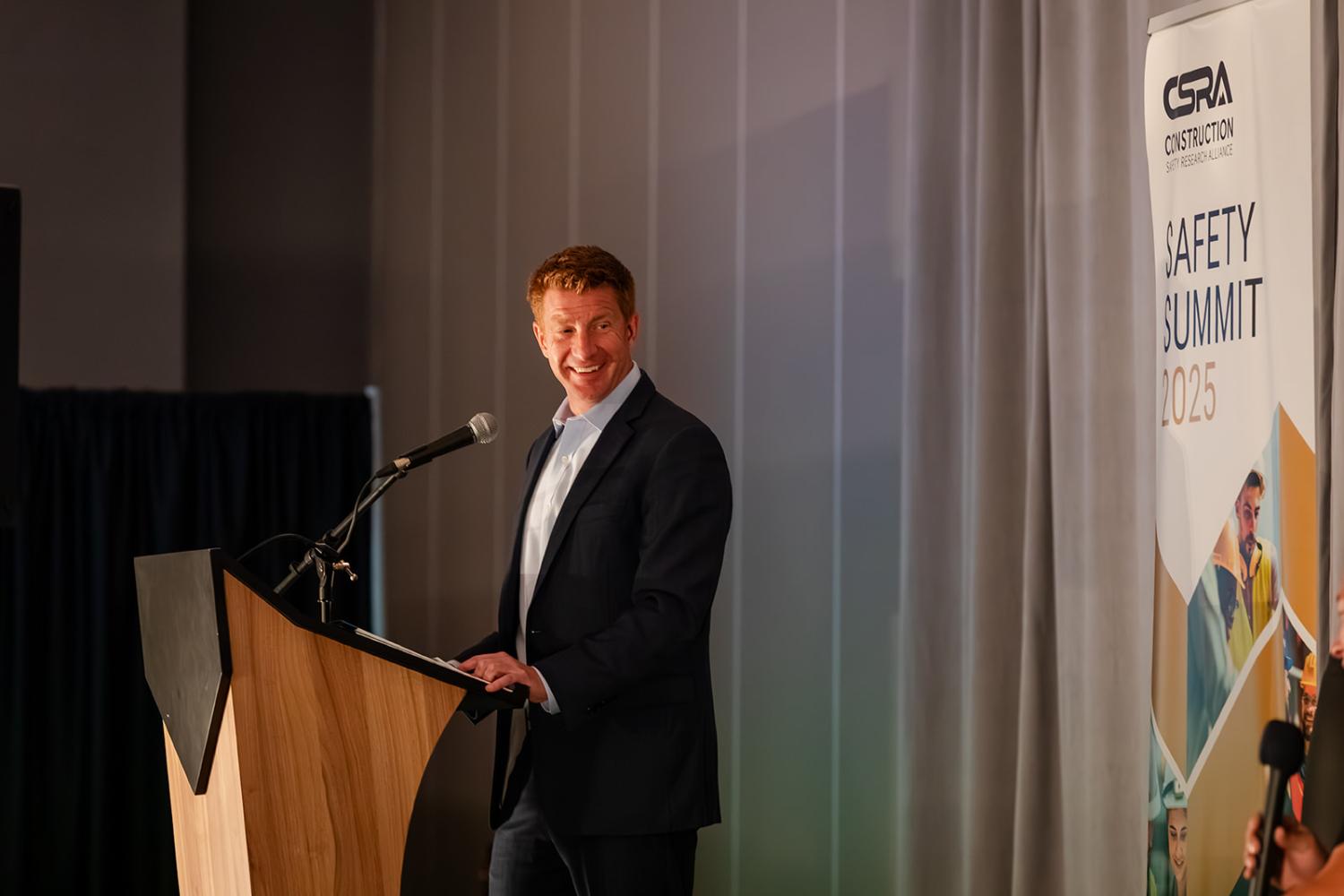Matt Hallowell in a suit stands at a podium smiling during a presentation at the CSRA Safety Summit 2025, with a banner for the event visible beside him.
