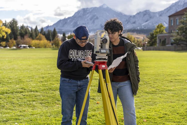 Two students by surveying equipment with the Flatirons in the background.