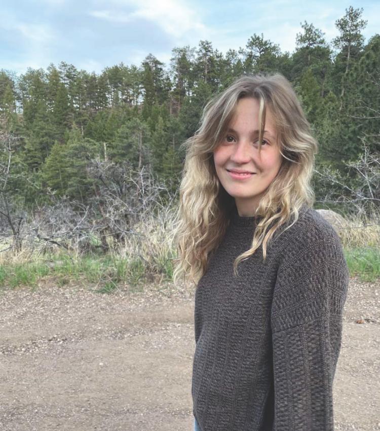 Macy Will standing in a sweater with some pine trees in the background.