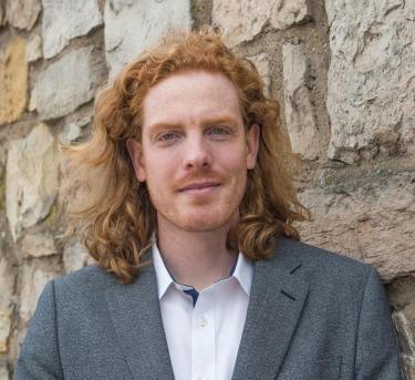 Jay Arehart in a suit jacket and white button-down sirt in front of a stone wall of a building