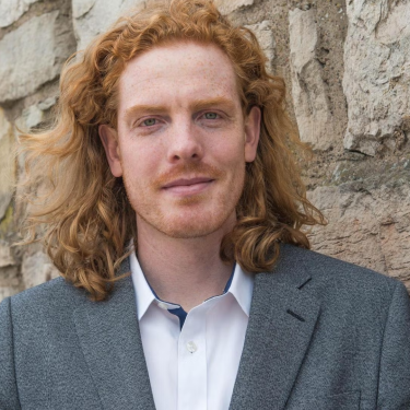 Jay Arehart in a white shirt and gray blazer in front of a stone wall.