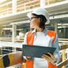A woman in a hard hat holding a clip board inside a building that is being built.