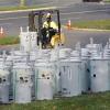 A worker in a high-visibility jacket and hard hat drives a forklift through rows of large cylindrical electrical transformers arranged outdoors on a paved lot. Traffic cones and a grassy area border the scene.