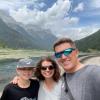 Amy Javernick-Will and her teenage child and husband casually dressed with a scenic mountain landscape and a river in the background. The father and son are wearing sunglasses and Amy has curly hair.