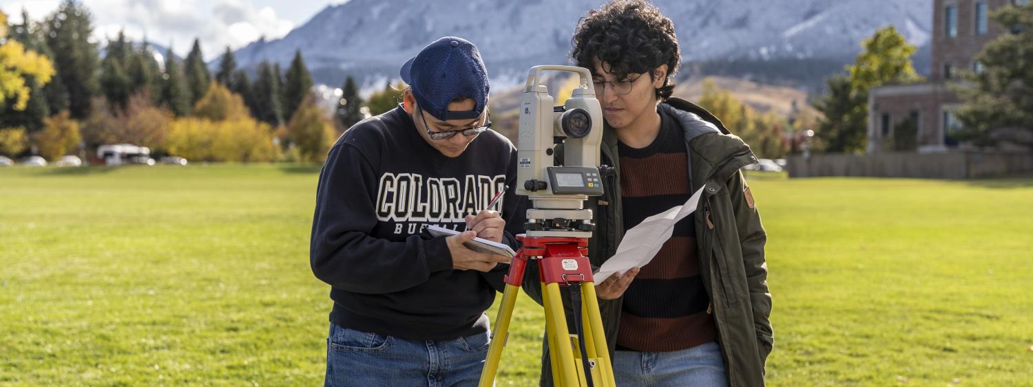 Two students by surveying equipment with the Flatirons in the background.